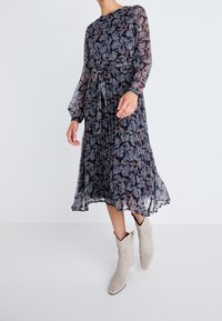 Blommönstrad, genomskinlig långärmad klänning med cinchad midja, flödande fåll och ett lätt tierat design. Bärs med ljusa ankelstövlar.