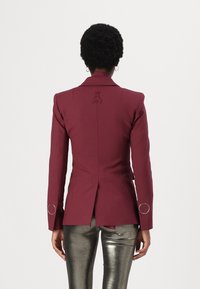 Blazer burdeos a medida con solapas pico, cierre de un solo botón, detalle bordado en la espalda y acentos circulares plateados en las mangas.