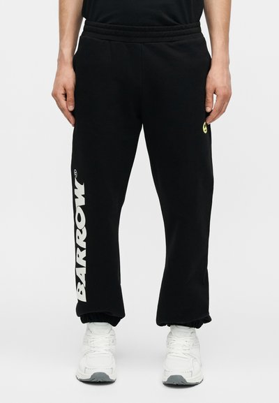 Άνδρας που φοράει μαύρη φόρμα με το κείμενο "BARROW" στο αριστερό πόδι και μικρό κίτρινο χαμογελαστό πρόσωπο στο δεξί μηρό, συνδυασμένη με λευκά αθλητικά παπούτσια.