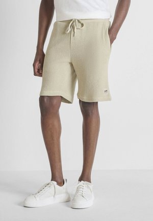 Hombre con pantalones cortos beige texturizados con cordón, camiseta blanca y zapatillas blancas, de pie sobre un fondo neutro.