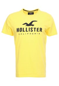 Gul bomullströja med rund halsringning, korta ärmar och en grafisk tryck med "HOLLISTER" och "CALIFORNIA" i mörkblått.