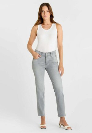 Donna in piedi che indossa un top bianco senza maniche, jeans grigio chiaro a vita alta e sandali bianchi con tacco e punta aperta su sfondo semplice.