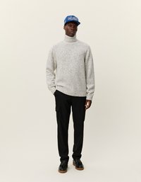 Hellgrauer Rollkragenpullover mit gesprenkelter Textur, kombiniert mit schwarzen Cargo-Hosen und einer blauen Mütze mit weißem Text. Schwarze Schuhe runden das Outfit ab.