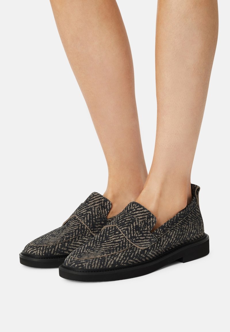 Pieds portant des mocassins slip-on noirs et gris à motifs avec semelles noires, montrés sur un fond uni clair.