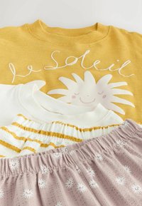 Chemise jaune avec un soleil souriant et le texte « le Soleil », chemise blanche à rayures jaunes, et pantalon rose à taille élastique avec motif marguerite blanc.