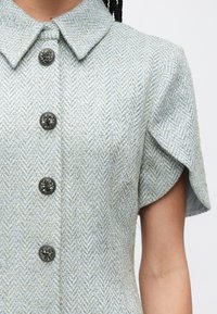 Kortärmad klänning i ljusblå fiskbensmönstrad tyg, med spetsig krage och utsmyckade svarta knappar längs fram.