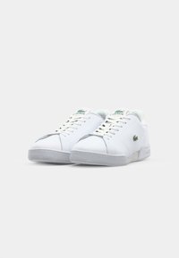 Ténis de couro branco com uma sola de borracha cinza texturizada, apresentando o logótipo verde da Lacoste e costura decorativa. Design com atacadores e bico redondo.