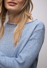 Femme aux longs cheveux blonds portant un pull bleu clair texturé, montrée des épaules aux lèvres sur un fond neutre.