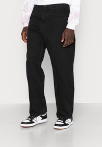 Black denim jeans with a straight-leg fit, featuring a mid-rise waist, five-pocket design, and contrasting stitching. Worn with black and white sneakers.