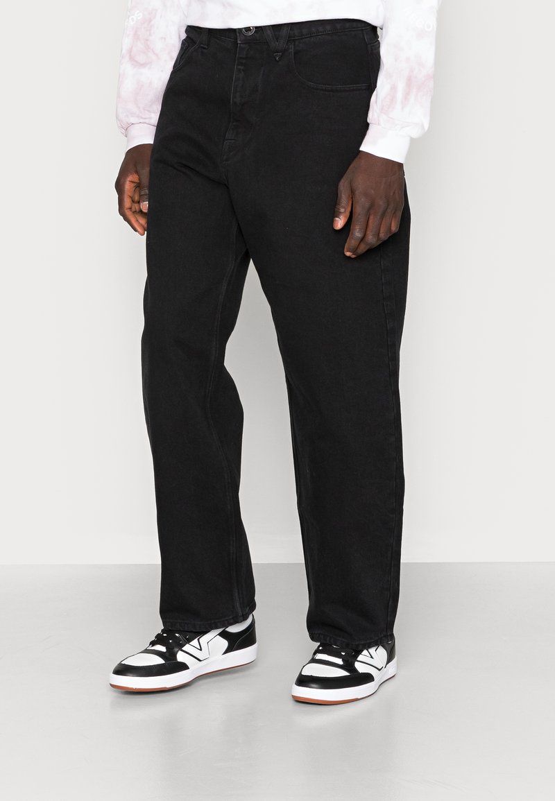Black denim jeans with a straight-leg fit, featuring a mid-rise waist, five-pocket design, and contrasting stitching. Worn with black and white sneakers.