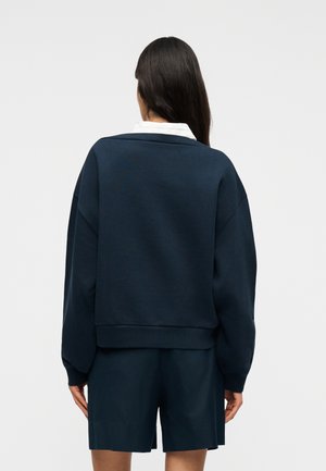 Γυναίκα με μακριά σκούρα μαλλιά που φοράει μπλε σκούρο φούτερ και ταιριαστό σορτς, στέκεται γυρισμένη προς τα πίσω σε απλό γκρι φόντο.