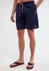 Tamsiai mėlynos maudymosi šortai su elastinga juostos, raudona ir balta virvele, bei šoninėmis kišenėmis. Apatinėje kojoje yra mažas baltas logotipas.