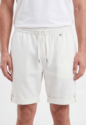 Hombre con pantalones cortos blancos con cordón y puños remangados, y una camiseta blanca lisa, de pie con los brazos relajados a los lados.