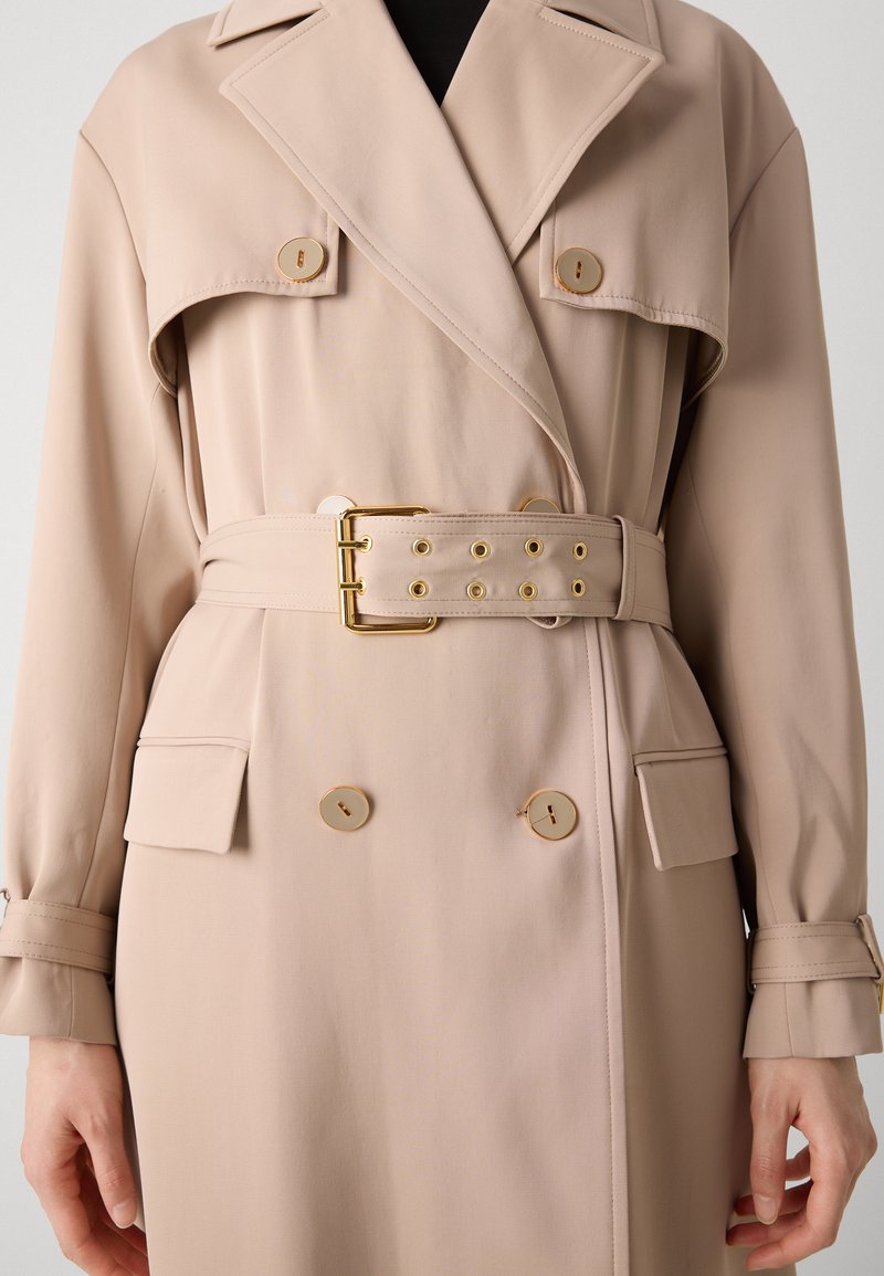 Beige Trenchcoat mit doppelter Knopfleiste, goldenen Hardware-Knöpfen, Stoffgürtel mit Ösen und zwei seitlichen Taschen.