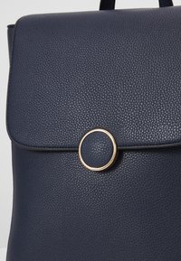 Mörkblå läderryggsäck med en strukturerad yta, som har ett runt guldspänne och en klaffstängning.