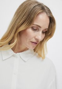 Blusa bianca con colletto realizzata in un tessuto testurizzato, caratterizzata da una chiusura con bottoni e una vestibilità morbida. Il colletto è morbido e con bordi arrotondati.