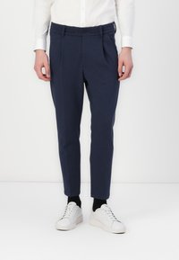 Pantalones ajustados color azul marino en tejido texturizado, con pliegues en la parte delantera, cierre de botón y bolsillos laterales, combinados con zapatillas blancas.