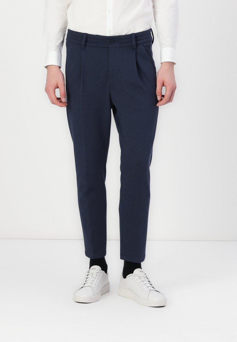 Pantalones ajustados color azul marino en tejido texturizado, con pliegues en la parte delantera, cierre de botón y bolsillos laterales, combinados con zapatillas blancas.