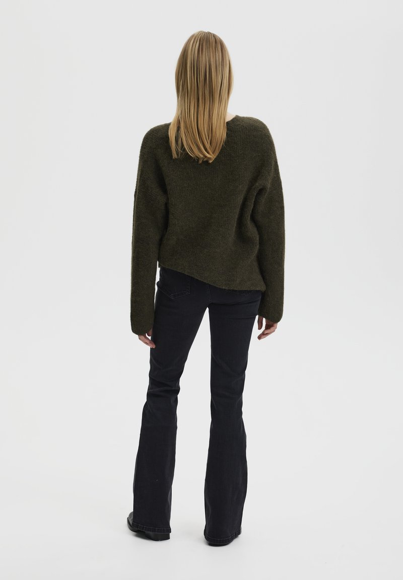 Suéter de gran tamaño en verde oscuro con un acabado texturizado, combinado con jeans negros de campana. Vista desde la parte posterior, mostrando el cabello largo y liso.