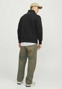 Schwarzer Pullover mit hohem Kragen, kombiniert mit grünlich-braunen Hosen. Verfügt über einen gerippten Saum und ein dezentes Logo-Detail auf der Rückseite.