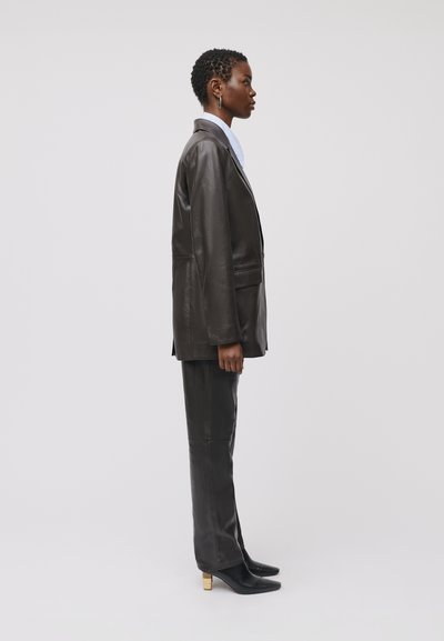 Blazer in pelle marrone scuro con spalle strutturate, tasche frontali, abbinato a pantaloni in pelle coordinati e tacchi a punta neri con dettagli dorati.