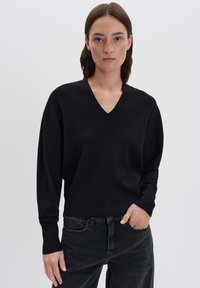 Frau mit glattem braunem Haar, die ein schwarzes V-Ausschnitt-langärmliges Oberteil und dunkle Jeans trägt und mit einer Hand in der Tasche steht.