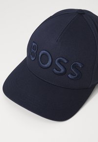 Marineblå caps med en strukturert design. Har et brodert "BOSS"-logo i en litt lysere nyanse. Laget av glatt stoff med en buet brem.