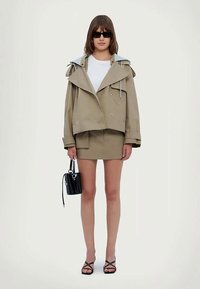 Beige, oversize dzseki széles gallérral és zsinóros kapucnival, egy illeszkedő bézs szoknyával és fekete táskával párosítva. A modell magassarkú szandálban áll.