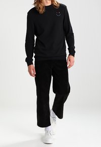 Person som bär en svart långärmad tröja med ett litet rött och blått smiley-ansikte, svarta vida byxor, vita strumpor och vita snörskor.