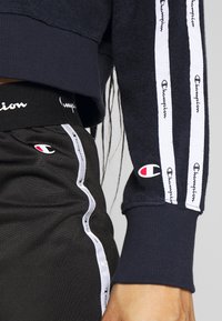 Pullover navy con polsini a costine, caratterizzato da strisce laterali bianche con il logo "Champion". Pantaloni corti neri con logo abbinato e bordature argentate.