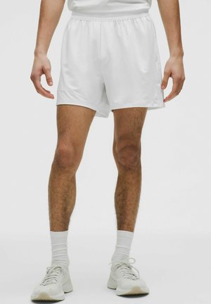 Homme portant un short de sport blanc, des chaussettes blanches et des baskets blanches, debout devant un fond uni.