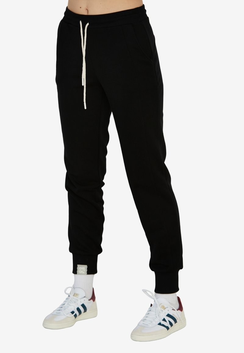 Czarne dresy z elastycznym paskiem w talii i kremowym sznurkiem; mają przednie kieszenie i ściągacze na końcu nogawek, zestawione z białymi butami i paskowanymi akcentami.