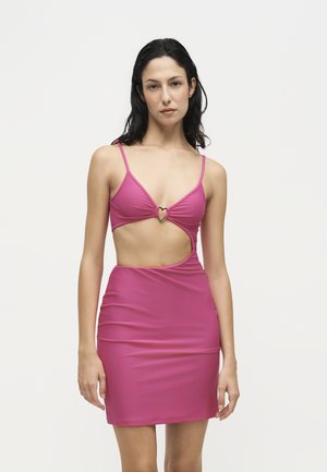 Femme aux cheveux foncés portant une mini-robe rose sans manches avec une découpe devant et un fermoir en forme de cœur, debout devant un fond blanc uni.