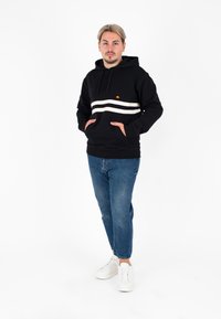 Felpa nera con strisce orizzontali bianche, tasche a marsupio e un piccolo logo sul petto, indossata con jeans blu e sneakers bianche.