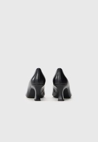Paire de chaussures en cuir noir à talon moyen vue de dos, posée sur un fond uni gris clair.