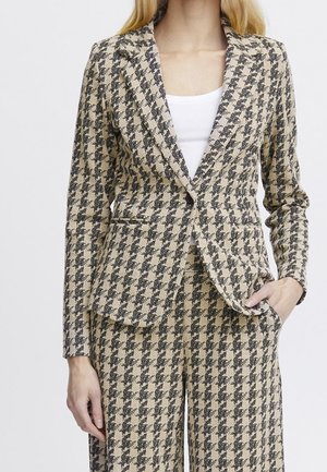 Kobieta w beżowo-czarnej marynarce w kratę i dopasowanych spodniach oraz białej bluzce, z jedną ręką w kieszeni, na jednolitym tle.