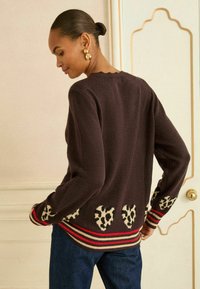 Brązowy sweter z sercami w wzór Leopard na dole, w paski w odcieniach czerwonym i beżowym na mankietach oraz ze scallopowym dekoltem. Noszony z ciemnymi dżinsami.