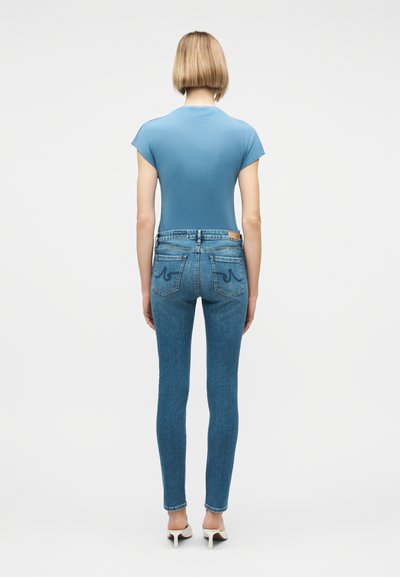 Γυναίκα με κοντά ξανθά μαλλιά που φοράει μπλε μπλούζα με κοντά μανίκια, εφαρμοστά μπλε τζιν με πίσω τσέπες και λευκά παπούτσια με τακούνι, που στέκεται με την πλάτη στραμμένη.