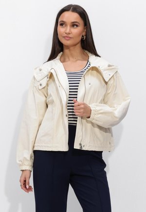 Vrouw draagt een crèmekleurig jack met rits over een zwart-wit gestreept topje en donkere broek, staand tegen een effen witte achtergrond.