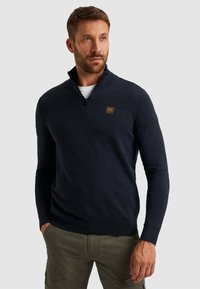 Marineblauwe gebreide pullover met een gestructureerd patroon, een ritskraag en een klein geel logo op de borst. Casual pasvorm met geribbelde boorden.