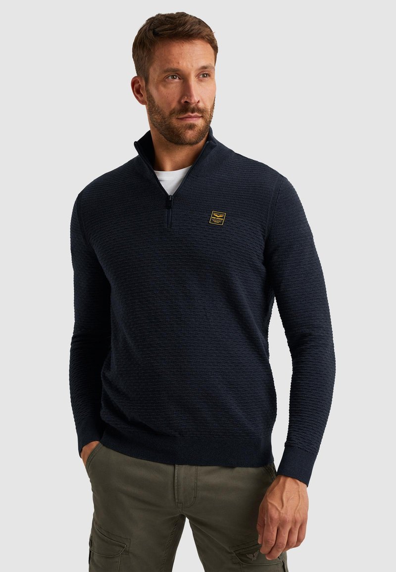 Marineblauwe gebreide pullover met een gestructureerd patroon, een ritskraag en een klein geel logo op de borst. Casual pasvorm met geribbelde boorden.