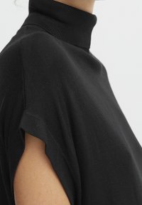 Svart polotröja med mjuk textur, korta ärmar med utskärningar och en åtsittande ribbad krage. Ren, minimalistisk design.