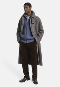 Ein Mann steht mit den Händen in den Taschen, trägt braune Kordhose, schwarze Schuhe, einen blauen Reißverschluss-Pullover und einen langen grauen karierten Mantel.