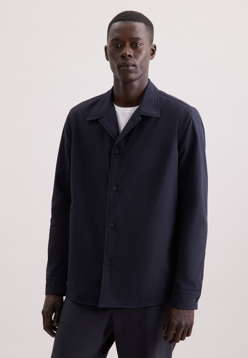 Granatowa koszula zapinana na guziki, lekki materiał, kołnierz rozłożony, długie rękawy, pojedyncza kieszeń na piersi, zaprojektowana z gładką teksturą i dopasowanym krojem.