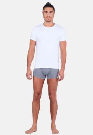 Uomo in piedi a piedi nudi che indossa una maglietta bianca aderente a maniche corte e boxer grigi su uno sfondo semplice.