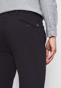 Ciemnogranatowe spodnie z kieszenią zapinaną na guzik, szlufkami na pasek i ozdobną metką. Otaczająca tkanina ma gładkie, teksturowane wykończenie.