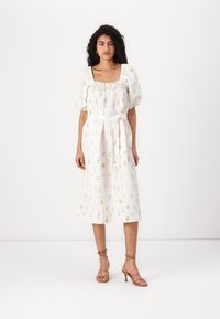 Vestido branco com estampado floral, decote quadrado elástico, mangas bufantes e cinto amarrado. Comprimento médio com um ajuste descontraído. Usado com sandálias de tiras.