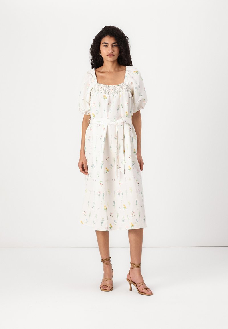 Vestido branco com estampado floral, decote quadrado elástico, mangas bufantes e cinto amarrado. Comprimento médio com um ajuste descontraído. Usado com sandálias de tiras.