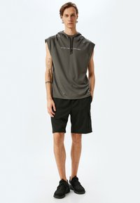 Ærmeløs grå hættebluse med hvid tekst, parret med sorte shorts med sidelommer og sorte sportsko med teksturerede såler.