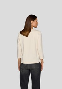 Helles beige Oberteil mit 3/4-Ärmeln und rundem Ausschnitt, aus einem weichen Stoff gefertigt, kombiniert mit dunkelgrauen Hosen, von hinten betrachtet.